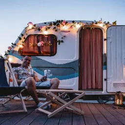 Uśmiechnięty mężczyzna siedzi na drewnianym krześle przed ozdobioną kwiatami i lampkami fotobudką w formie przyczepy kempingowej, trzymając w ręku pasek zdjęć.