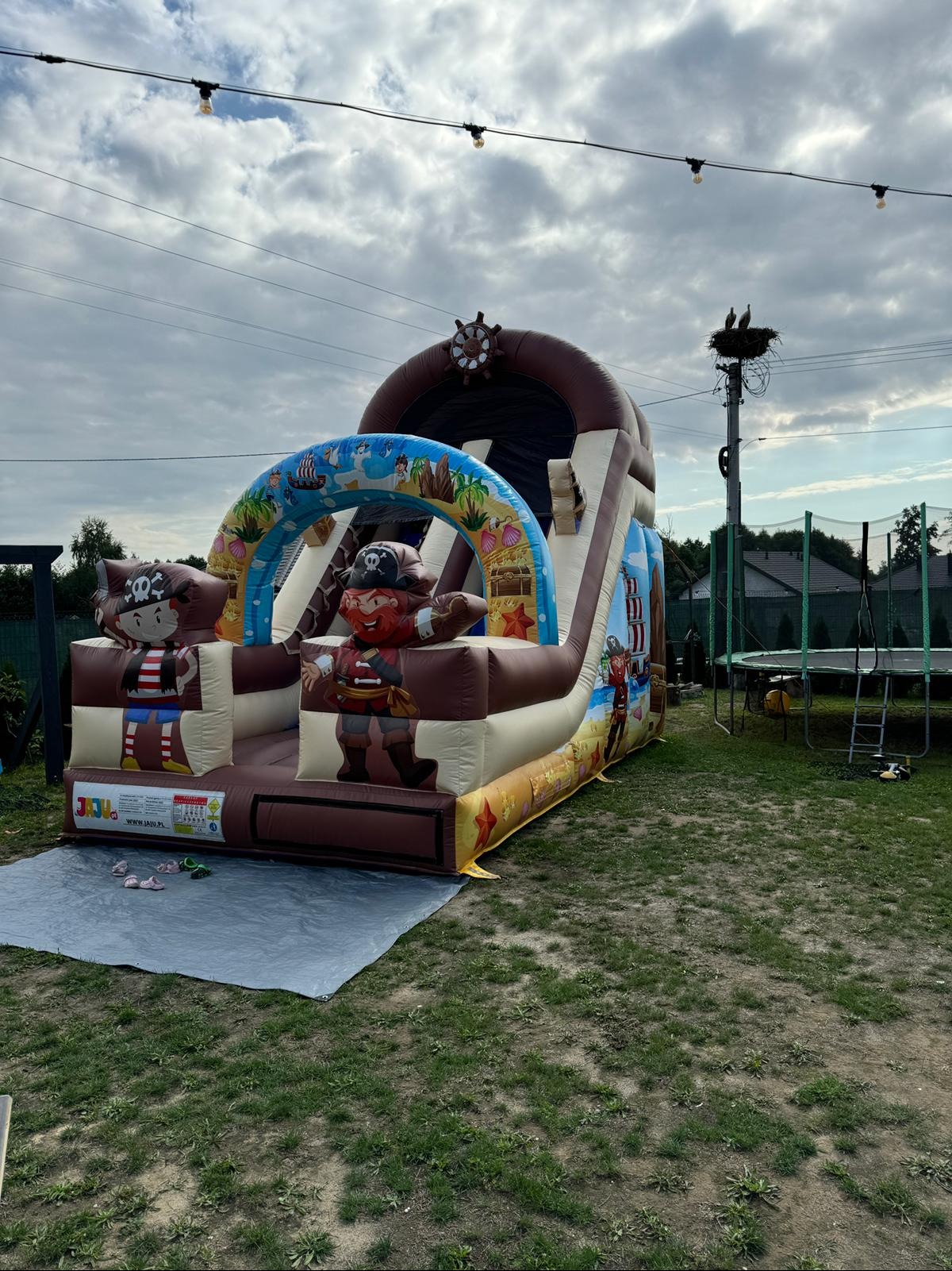 Piracki dmuchany zamek ze zjeżdżalnią, rozstawiony na trawie w Czarnej, z motywami piratów, palm i skrzyń ze skarbami. W tle widoczny słup z gniazdem ptaków i trampolina.