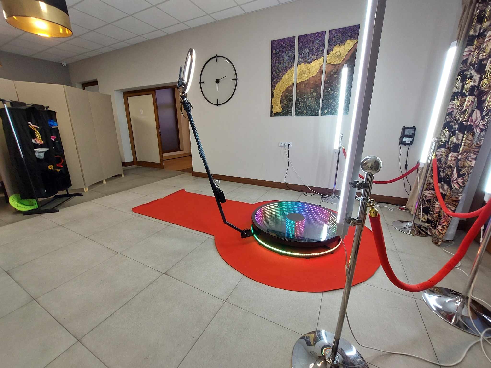 Okrągła platforma z kolorowym oświetleniem LED na czerwonym dywanie, otoczona słupkami z czerwonym sznurem, w tle ściana z obrazami i zegarem.