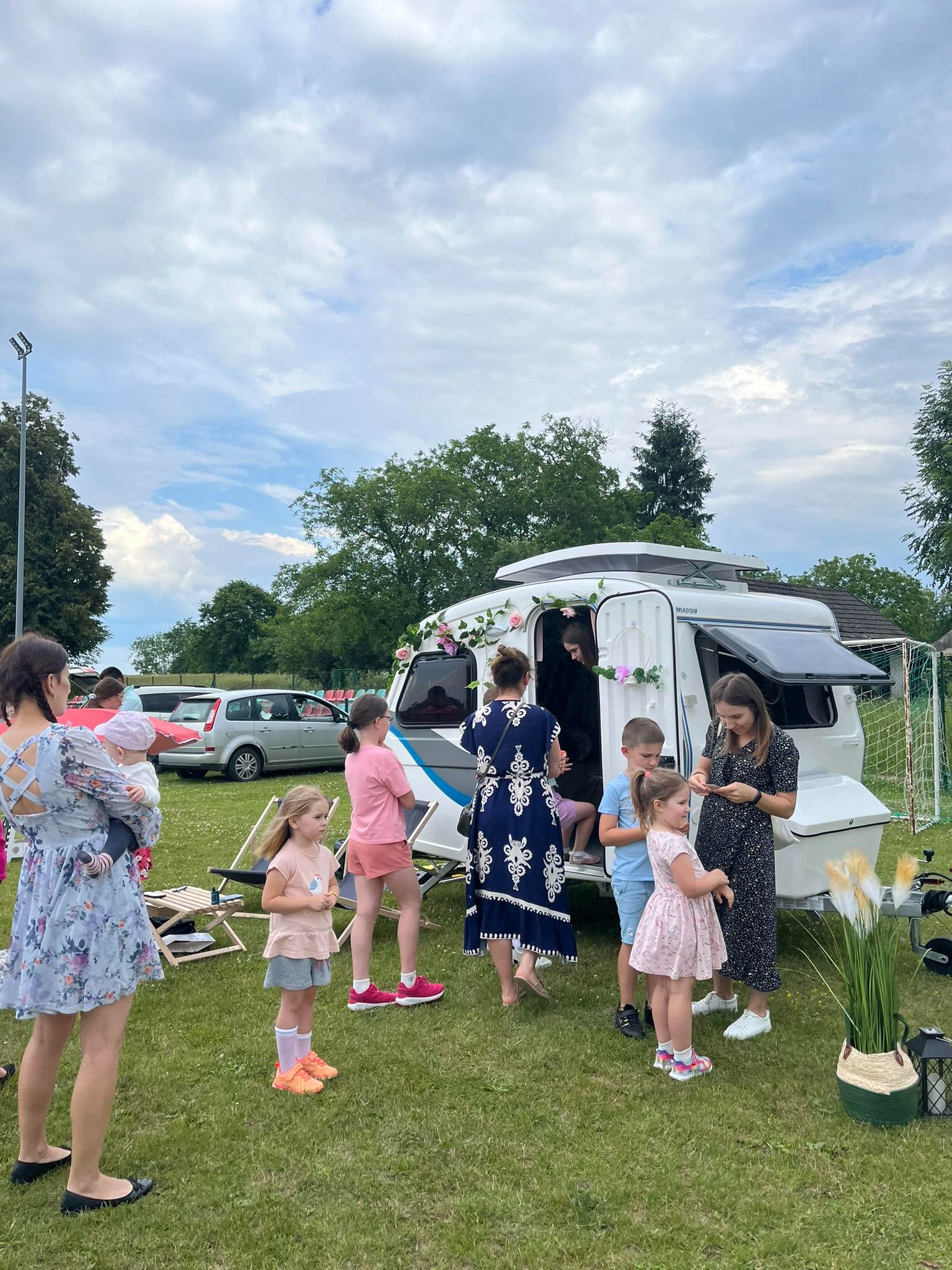 Udekorowana kwiatami fotobudka w formie przyczepy kempingowej otoczona grupą dzieci i dorosłych na zielonym trawniku podczas imprezy plenerowej.