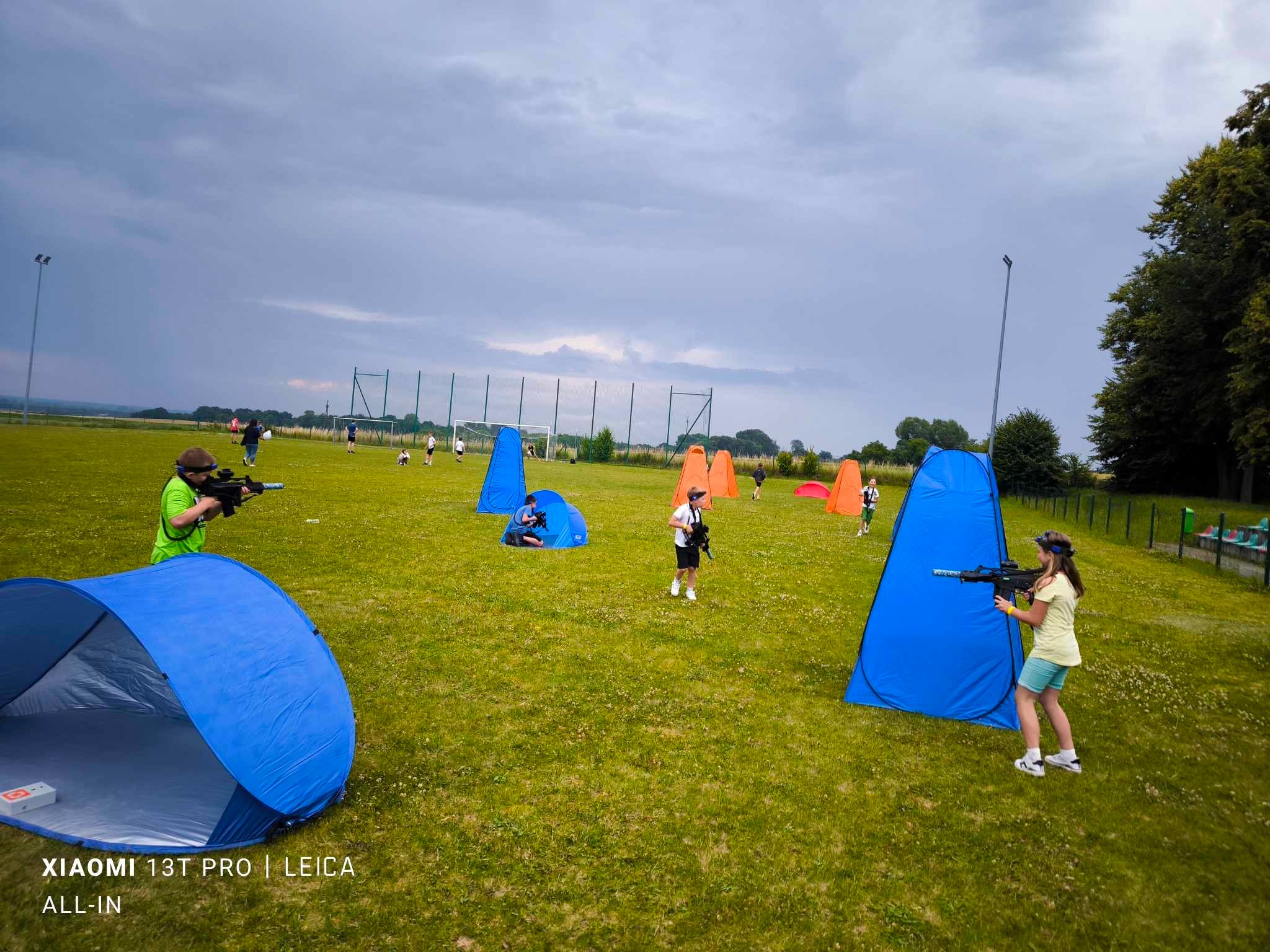Grupa dzieci i dorosłych grających w laserowy paintball na trawiastym terenie z niebieskimi i pomarańczowymi namiotami; pochmurne niebo w tle.