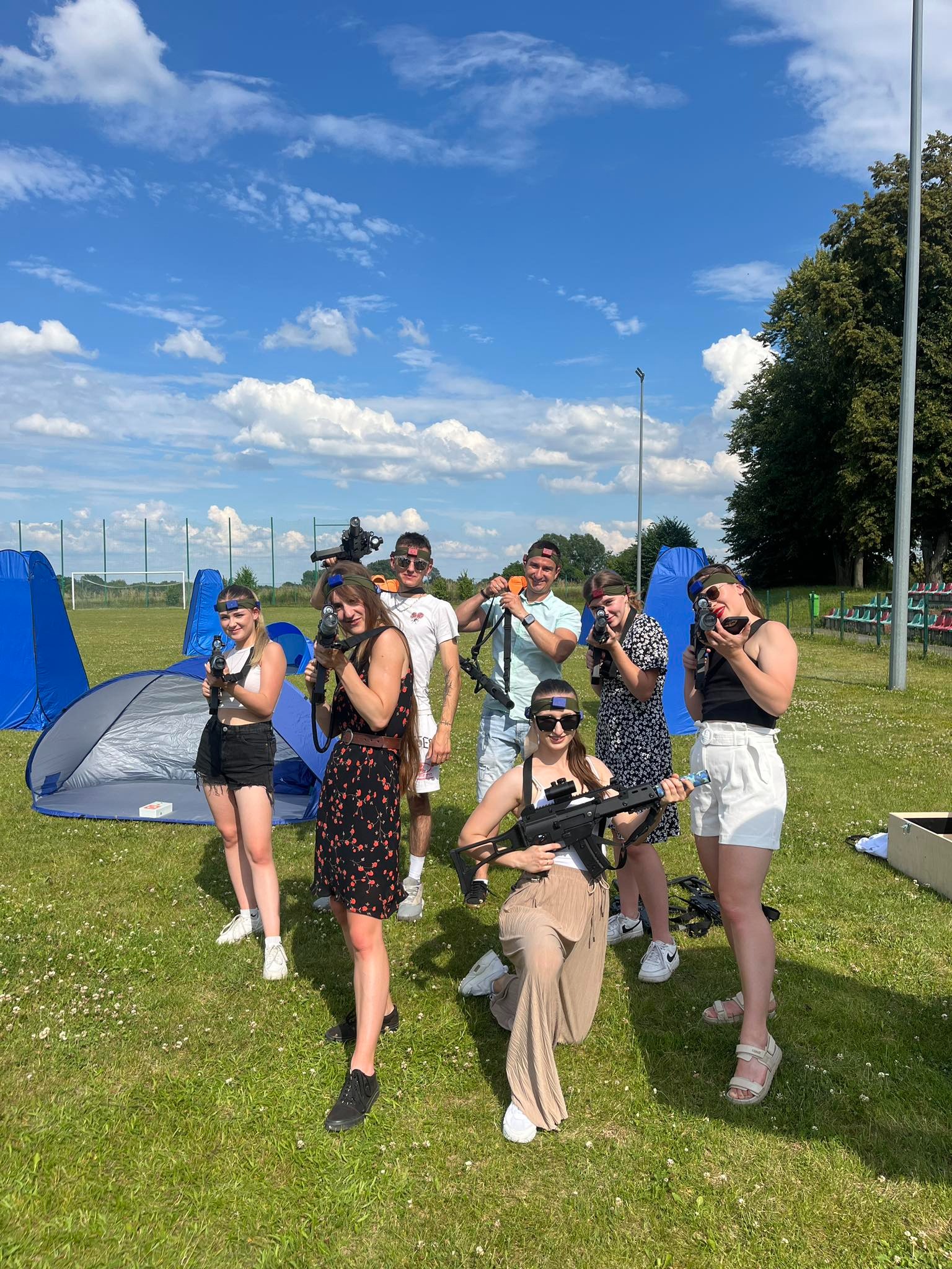 Grupa młodych ludzi, w okularach ochronnych i z laserowymi pistoletami, pozuje na trawiastym terenie z namiotami i trybunami w tle, w słoneczny dzień.