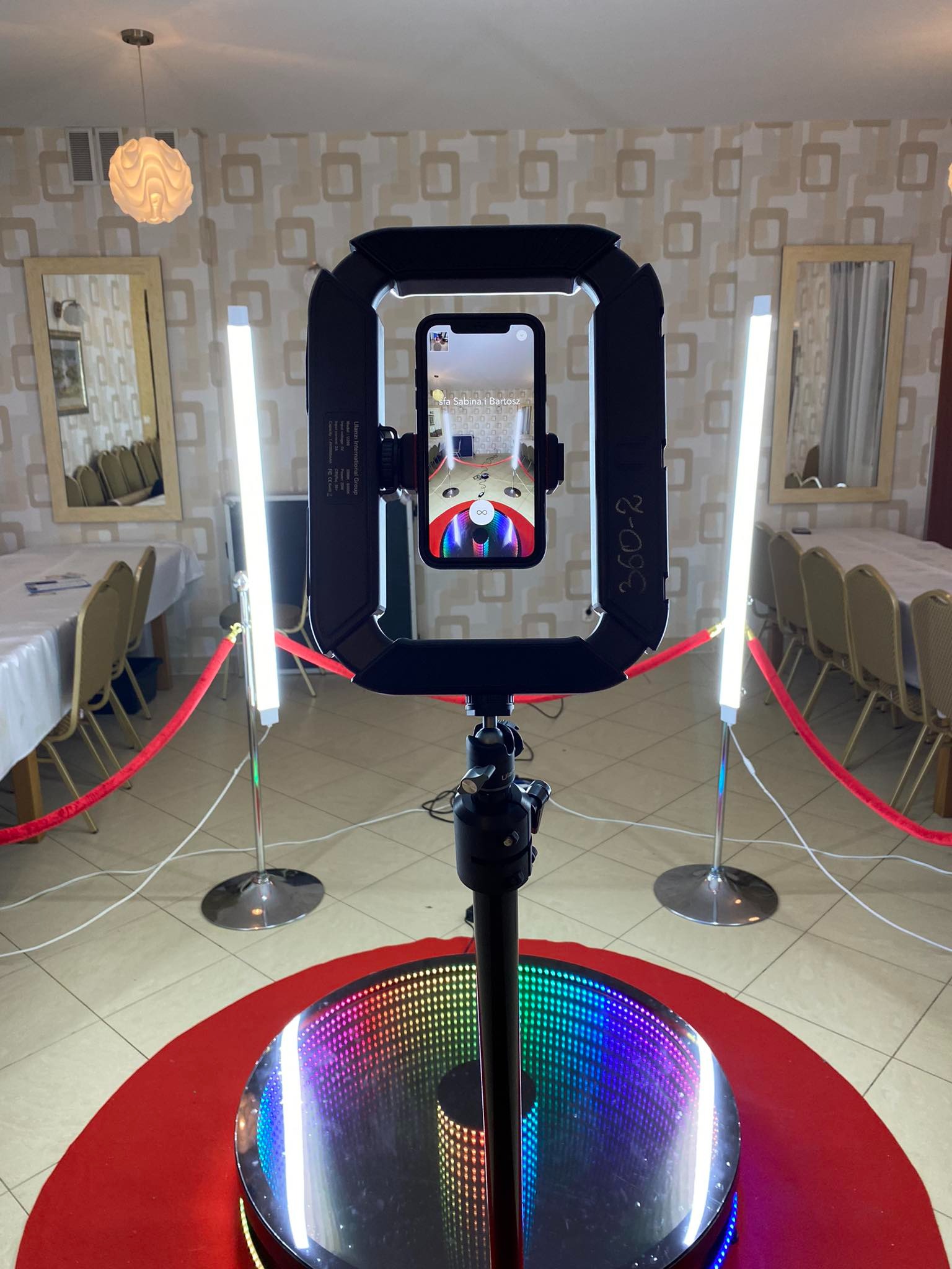 Automatyczna platforma obrotowa 360 stopni z oświetleniem LED i uchwytem na smartfon, ustawiona na czerwonym dywanie w sali bankietowej z nakrytymi stołami.