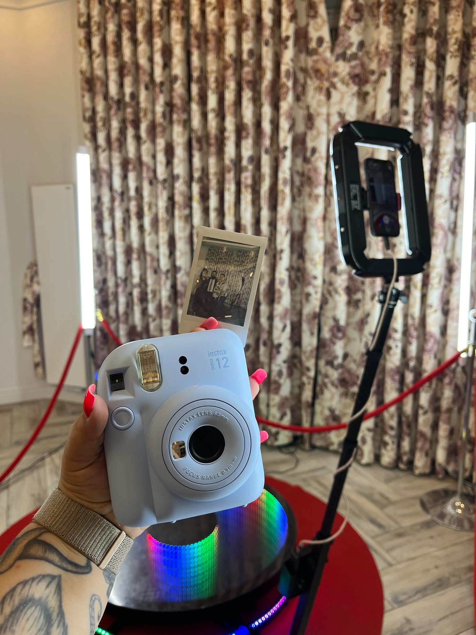 Osoba trzyma w dłoni aparat Instax mini 12 z wydrukowanym zdjęciem, w tle widoczna lampa pierścieniowa i dekoracyjne zasłony.