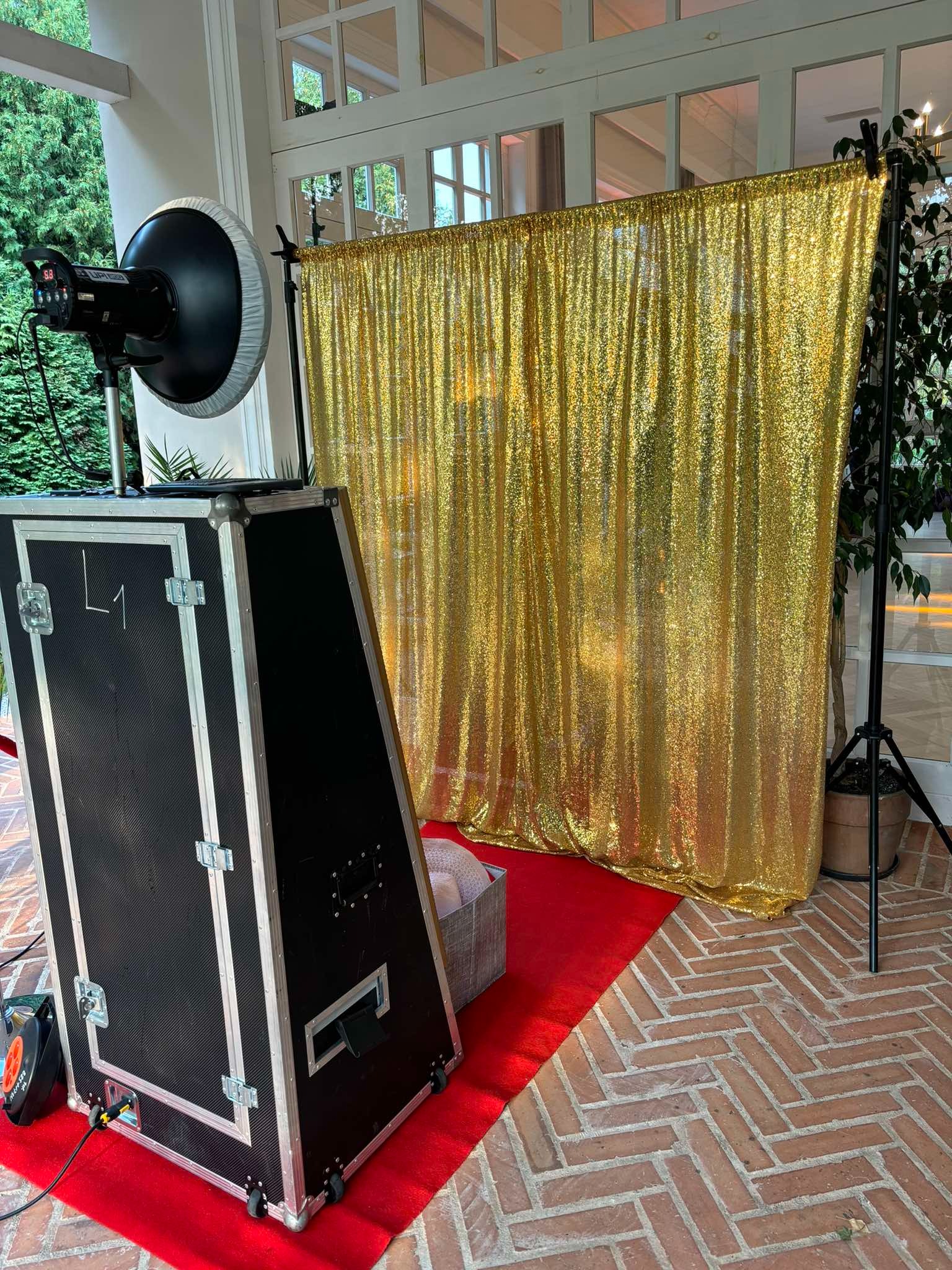 Czarna fotobudka z lampą studyjną i złotym, błyszczącym tłem na czerwonym dywanie, ustawiona na ceglanej podłodze w jodełkę.
