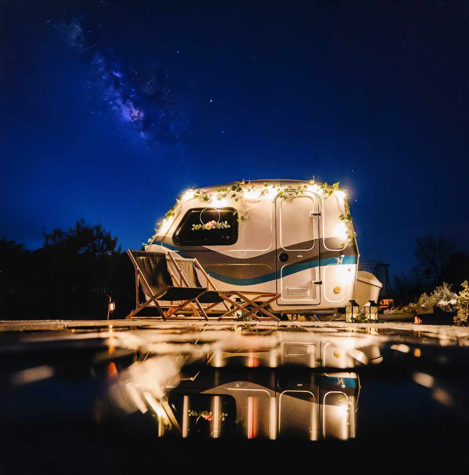 Przyczepa kempingowa udekorowana światełkami i kwiatami, z dwoma krzesłami plażowymi przed nią, nocne niebo z Drogą Mleczną w tle, odbicie w kałuży na pierwszym planie.