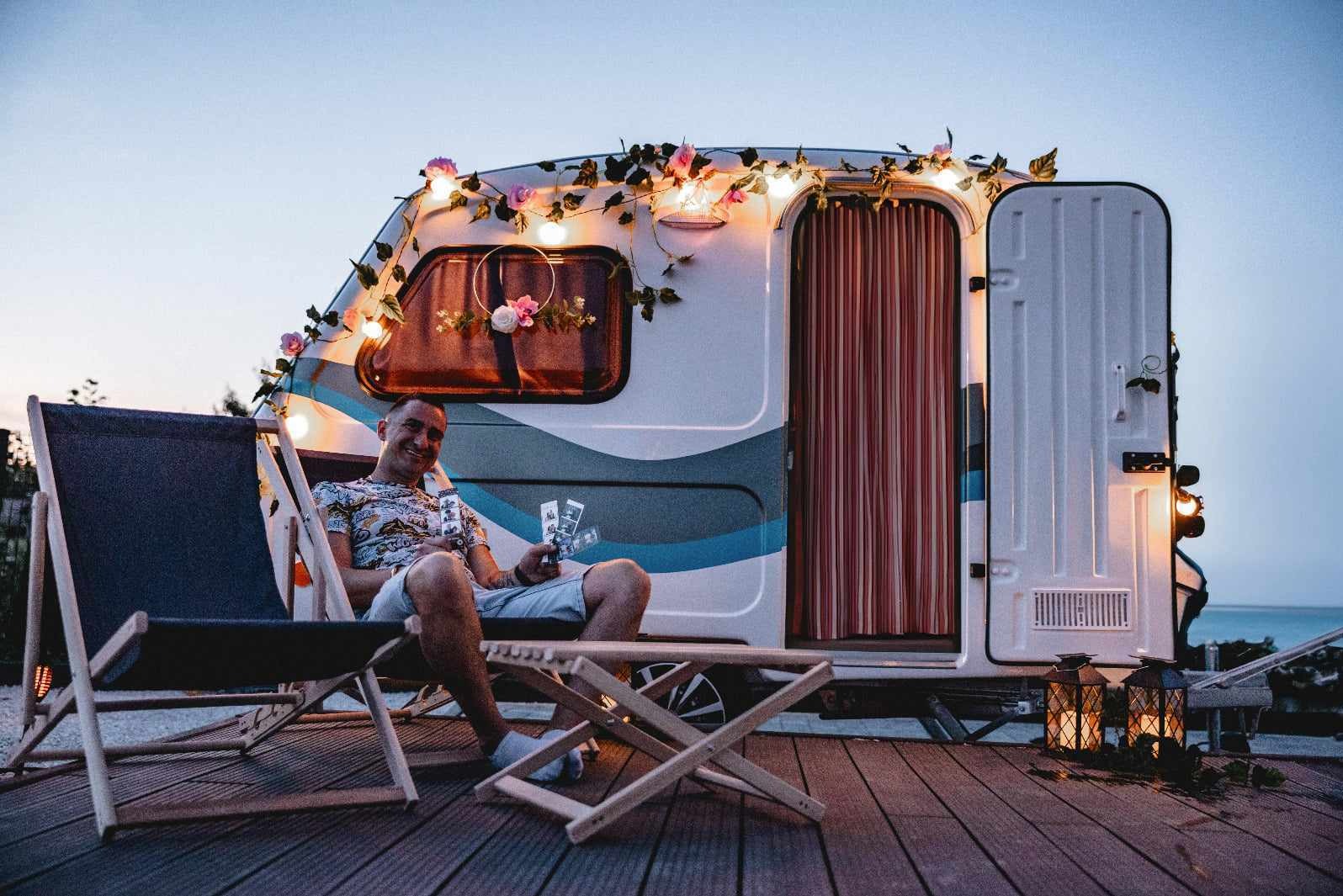 Uśmiechnięty mężczyzna siedzi na drewnianym krześle przed ozdobioną kwiatami i lampkami fotobudką w formie przyczepy kempingowej, trzymając w ręku pasek zdjęć.