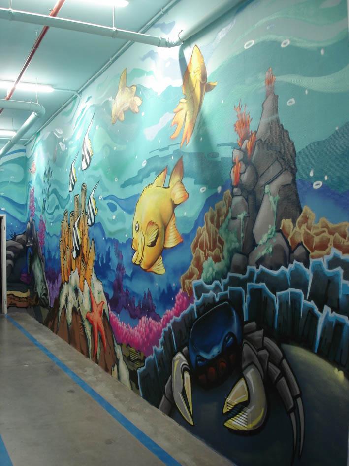Iluzjonistyczne malowidło ścienne przedstawiające podwodny świat z rybami, koralowcami i krabem w realistycznym stylu, biegnące wzdłuż korytarza z niebieską linią podłogową.