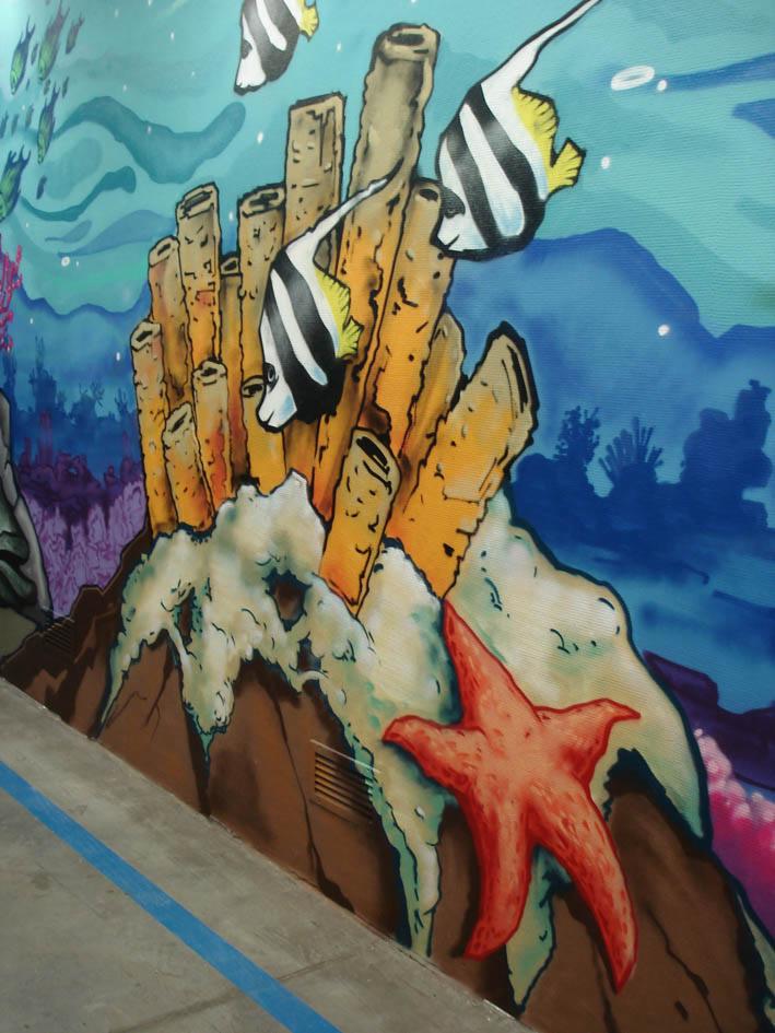 Ściana z muralem przedstawiającym podwodny świat z rybami żaglowymi, koralowcami i rozgwiazdą, namalowanym przy podłodze z niebieską linią.