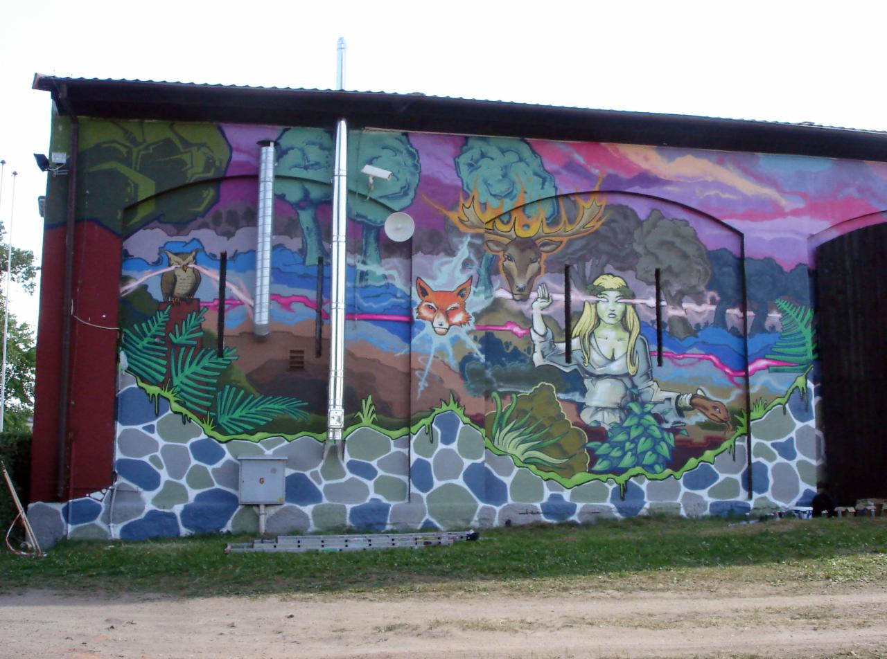 Duży mural na ścianie budynku przedstawiający fantastyczny krajobraz z sową, lisem, jeleniem, kobietą z promieniami w oczach i roślinami, z widocznymi elementami infrastruktury budynku.