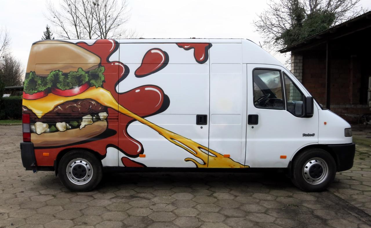Biały samochód dostawczy z grafiką burgera i keczupu, wykonaną metodą malowania artystycznego, zaparkowany na brukowanym podwórku.