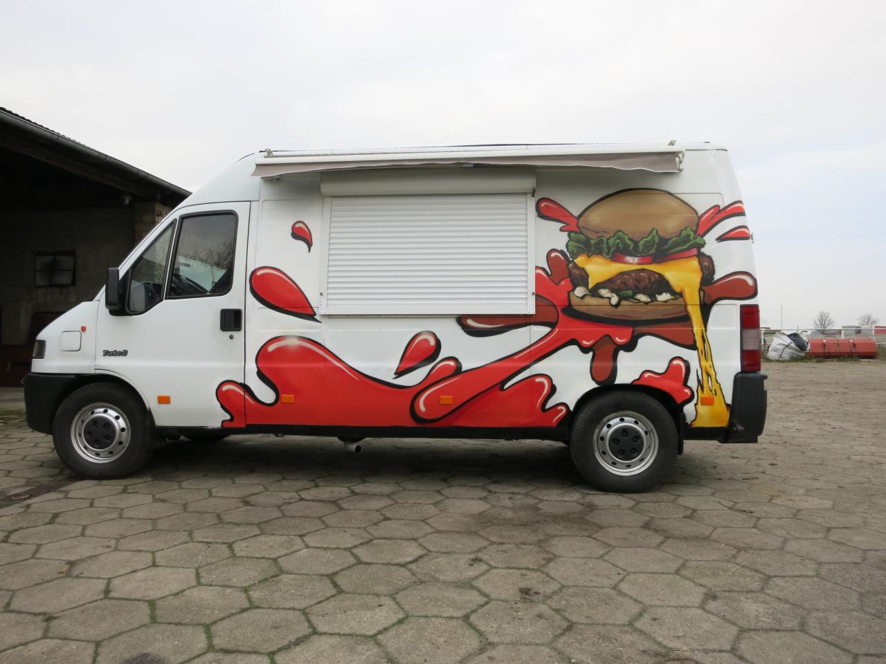 Biały food truck z pomalowaną grafiką burgera i czerwonymi plamami imitującymi sos, zaparkowany na betonowym placu.