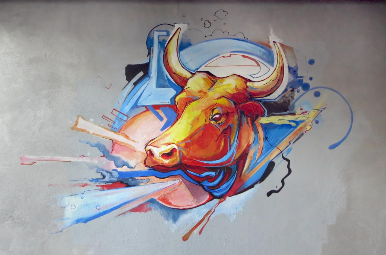 Abstrakcyjne malowidło ścienne przedstawiające głowę byka w odcieniach pomarańczy i żółci, otoczoną geometrycznymi kształtami w kolorach niebieskim i szarym, na szarym, teksturowanym tle.