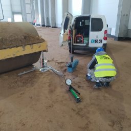 Centrum Geologii i Geotechniki Sp. z o.o. - Pracownik w kamizelce mierzy zagęszczenie gruntu ubijarką dynamiczną, obok walec i samochód z logo firmy. Hala z betonowymi kolumnami w tle.