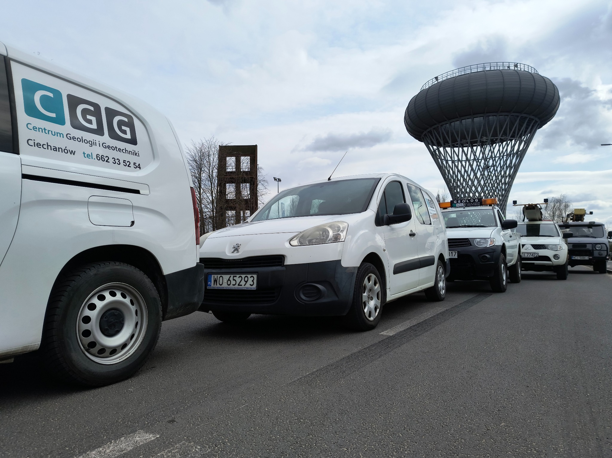Flota aut Centrum Geologii i Geotechniki z Ciechanowa zaparkowana na asfalcie. W tle wieża ciśnień i konstrukcja. Na pierwszym planie biały Peugeot z numerem WO 65293.