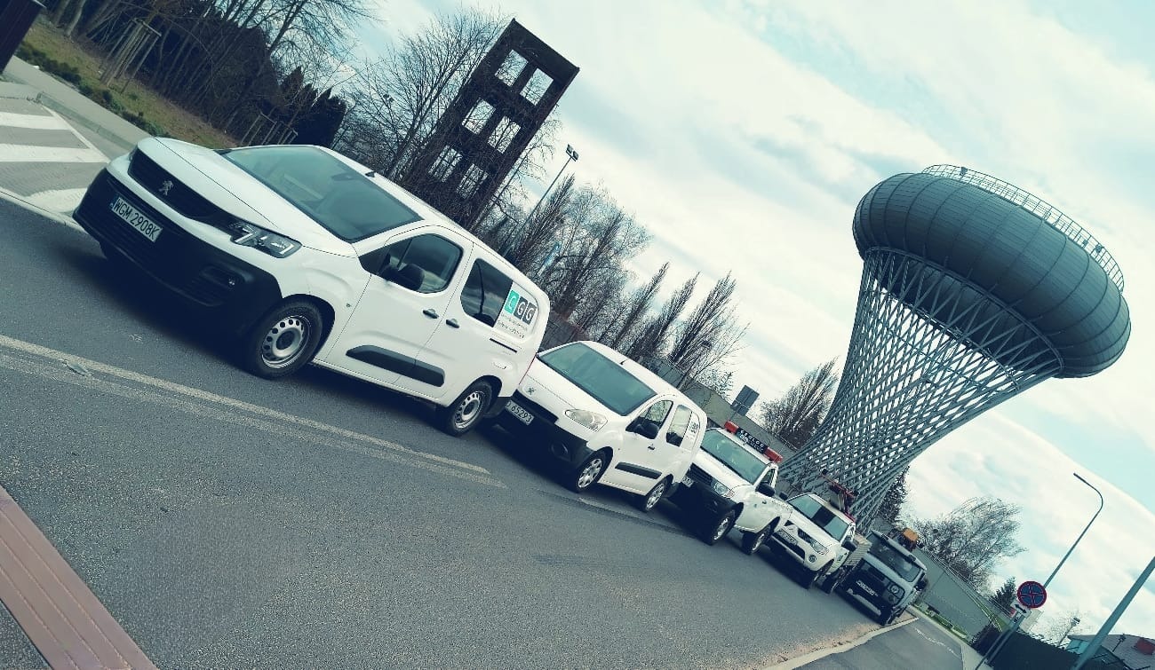 Flota białych samochodów firmy geologicznej ustawiona w rzędzie na tle wieży ciśnień i szarego nieba. Uwagę zwraca logo na pierwszym samochodzie.