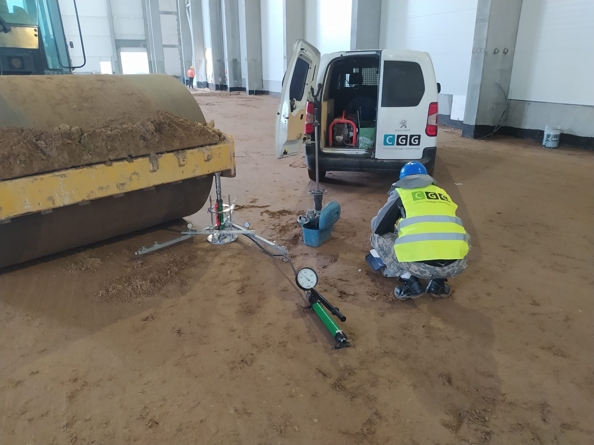 Pracownik w kamizelce mierzy zagęszczenie gruntu ubijarką dynamiczną, obok walec i samochód z logo firmy. Hala z betonowymi kolumnami w tle.
