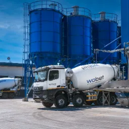 Dwa betonowozy firmy WOBET na tle silosów i niebieskiego budynku betoniarni w słoneczny dzień.