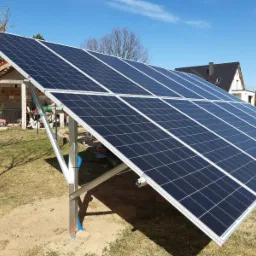 Instalacja fotowoltaiczna na metalowej konstrukcji, widoczna na tle trawnika i budynków mieszkalnych pod błękitnym niebem.