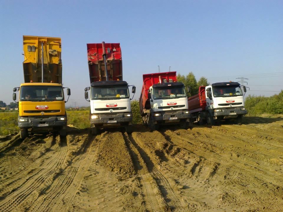 Cztery ciężarówki wywrotki, żółta i białe z czerwonymi skrzyniami, stoją na ziemi z podniesionymi skrzyniami ładunkowymi, widoczne ślady opon na piasku.