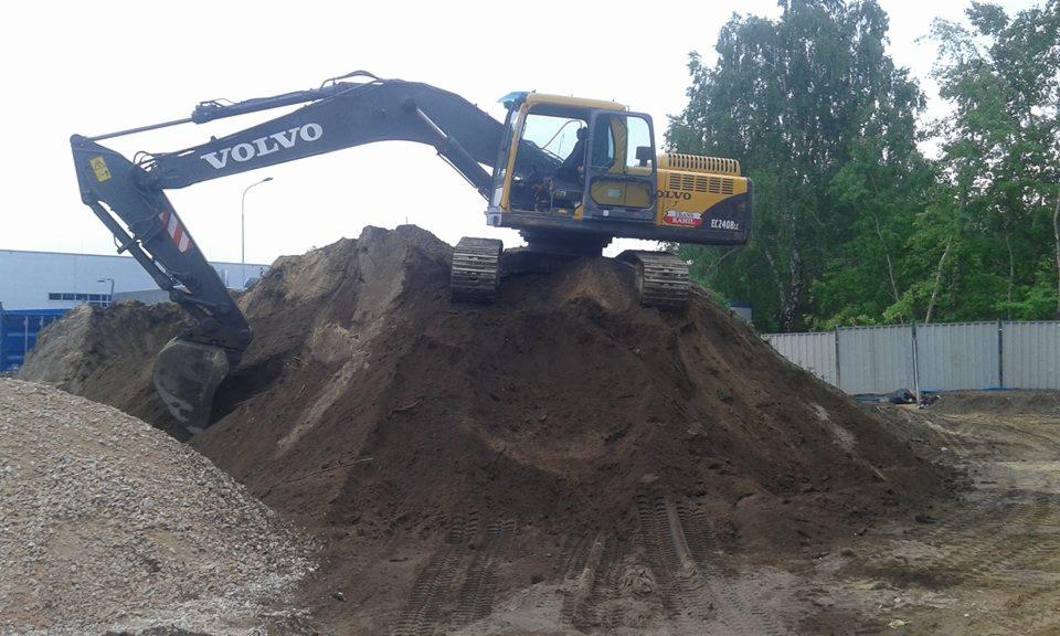 Żółto-czarna koparka Volvo EC240B LC na szczycie hałdy ziemi, widoczne ślady opon na ziemi, w tle budynek przemysłowy i drzewa.