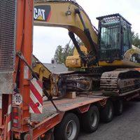 Żółta koparka CAT zabezpieczona pasami na niskopodwoziowej platformie transportowej w Brzezinach, widok z boku na załadunek z elementami odblaskowymi i tablicą ograniczenia prędkości.