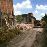 Żółta koparka Volvo usuwa gruz ceglany po rozbiórce budynku, widoczne fragmenty muru i piach, w tle budynek z czerwonej cegły i drzewa.