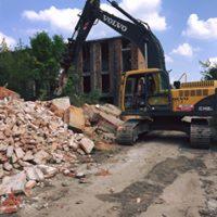 Żółta koparka Volvo z czarnym ramieniem, usuwająca gruz ceglany z terenu budowy. W tle widoczny budynek w trakcie rozbiórki i błękitne niebo z chmurami.