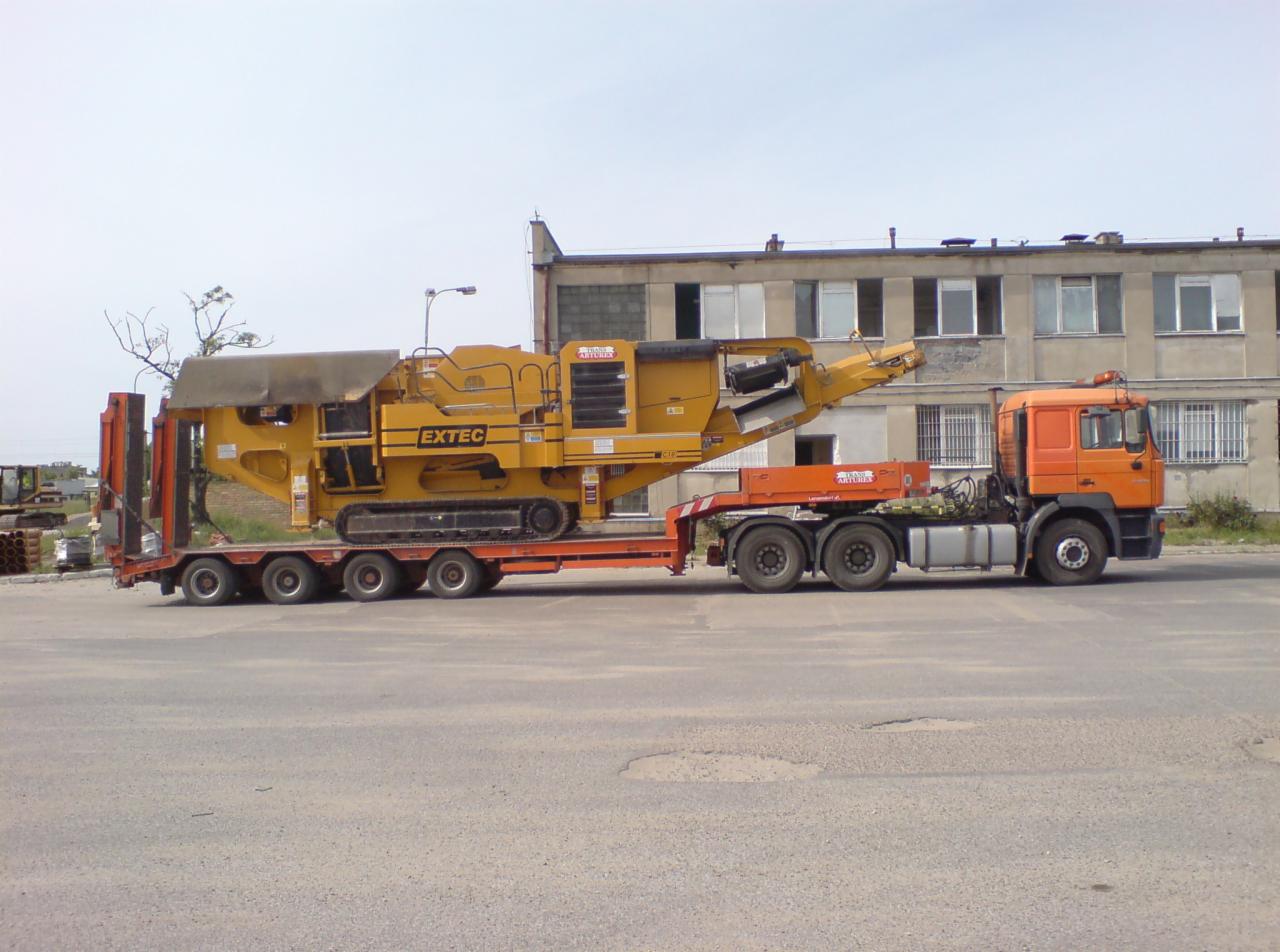 Pomarańczowa ciężarówka transportuje żółtą maszynę do kruszenia kamieni marki Extec na niskopodwoziowej platformie, na tle szarego budynku mieszkalnego.