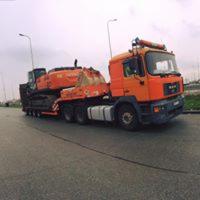 Pomarańczowy ciągnik siodłowy z naczepą niskopodwoziową przewożący pomarańczową koparkę gąsienicową na tle szarej drogi i pochmurnego nieba.