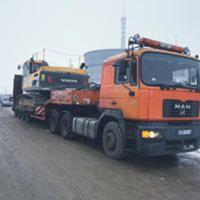 Pomarańczowa ciężarówka MAN z niskopodwoziową naczepą transportuje żółtą koparkę VOLVO. Widoczne elementy mocujące ładunek. W tle szara, zimowa sceneria i komin przemysłowy.