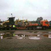 Pomarańczowy samochód ciężarowy przewozi na platformie ciężki sprzęt budowlany, prawdopodobnie równiarkę, zabezpieczoną pasami transportowymi, na placu z kałużami.