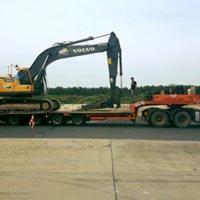 Żółto-czarna koparka Volvo na niskopodwoziowej platformie transportowej, zabezpieczona pasami, widoczny operator i otwarty teren w tle.