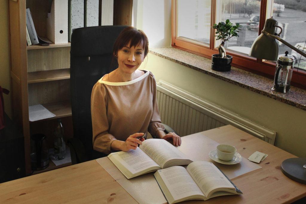 Dietetyczka uśmiecha się siedząc przy biurku z otwartymi książkami, filiżanką herbaty i lampką w stylu vintage, bonsai w tle przy oknie.