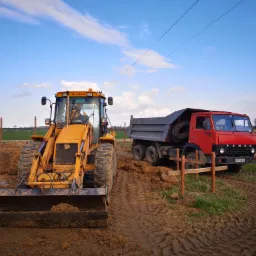 Żółta koparko-ładowarka JCB z operatorem i czerwona ciężarówka KAMAZ na placu budowy z widocznymi śladami opon w błocie, w tle słupy i druty wysokiego napięcia pod błękitnym niebem.