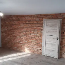 Wnętrze pokoju z wyeksponowaną ścianą z czerwonej cegły i jasnymi, drewnopodobnymi panelami podłogowymi, uzupełnione nowoczesnymi drzwiami wewnętrznymi z poziomymi żłobieniami i czarną klamką.