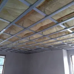 Montaż metalowego stelażu sufitu podwieszanego z widoczną wełną mineralną izolacyjną, surowe ściany w tle, okno z widokiem na zieleń.