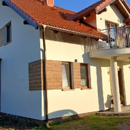 Jasna elewacja domu jednorodzinnego z drewnianymi okiennicami, balkonem i zadaszeniem tarasu z przezroczystymi ścianami.