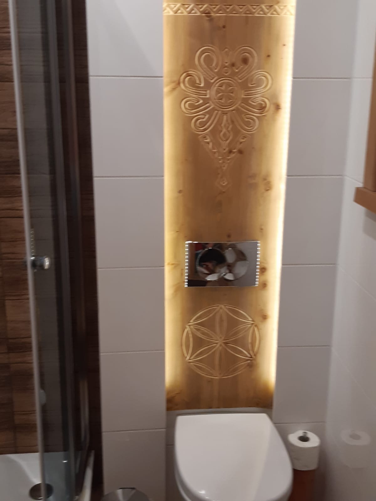 Oryginalna aranżacja toalety z drewnianym panelem zdobionym ornamentami, podświetlonym od tyłu, zamontowanym na ścianie z białych płytek.