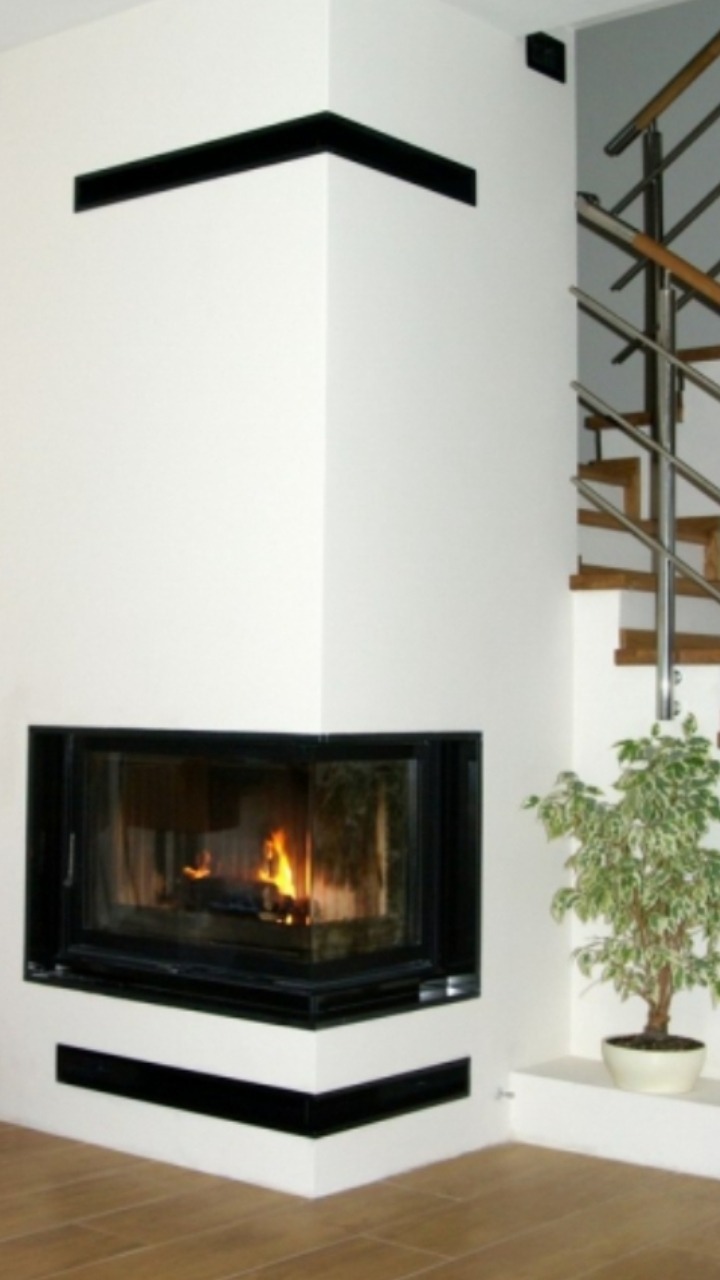 Wnętrze salonu z narożnym kominkiem z palącym się ogniem, drewnianą podłogą, białymi ścianami z czarnymi akcentami, schodami z metalową balustradą i zielonym drzewkiem w doniczce.