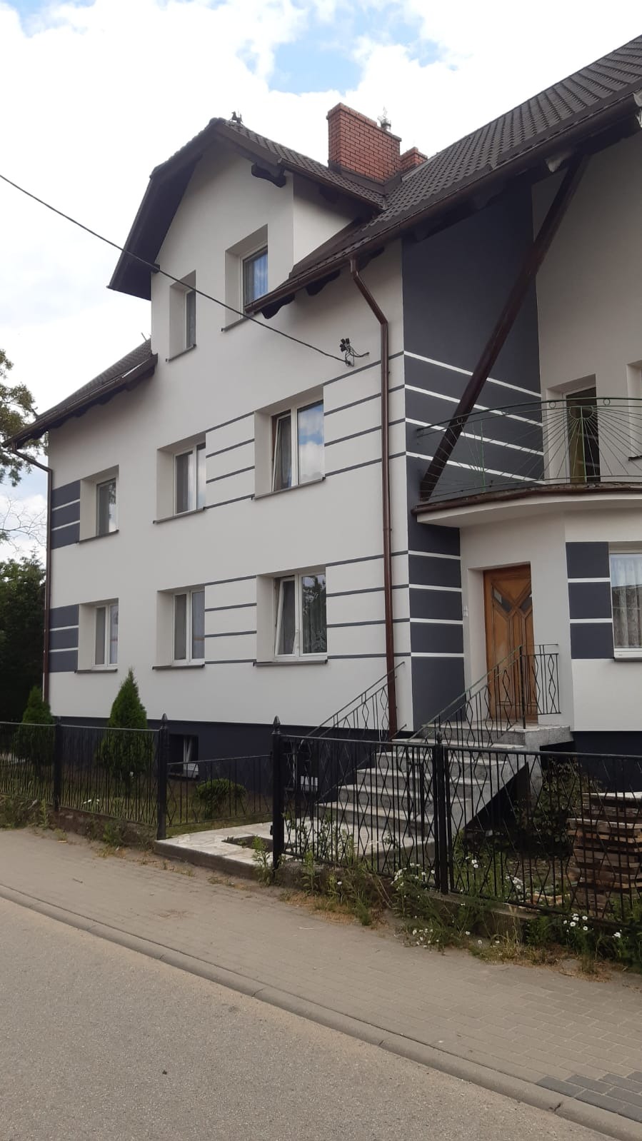 Dwupiętrowy dom z nową, biało-szarą elewacją, widoczne poziome pasy i drewniane drzwi wejściowe, czarna metalowa balustrada przy schodach.