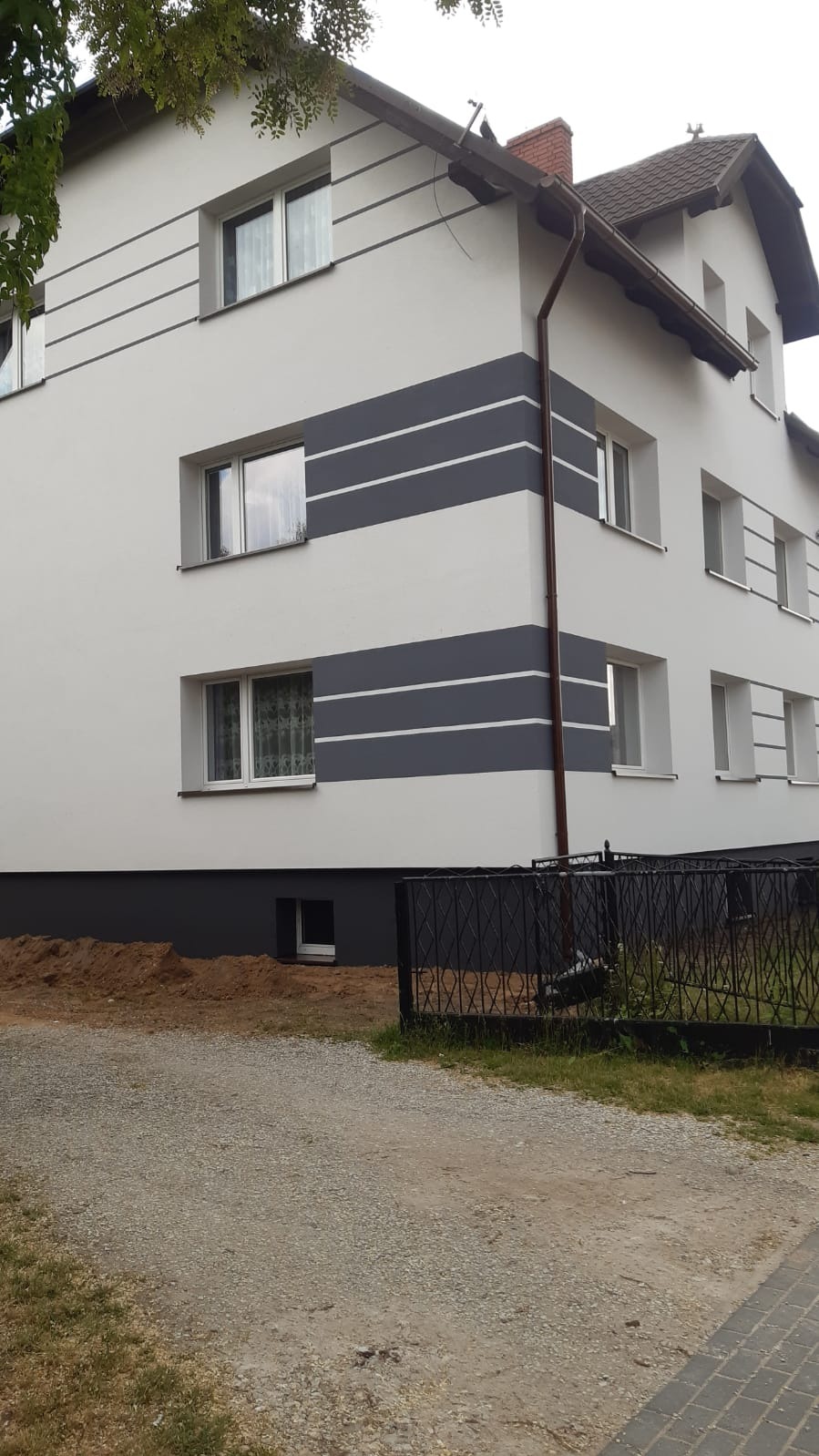 Dwukondygnacyjny dom z jasną elewacją i szarymi pasami dekoracyjnymi, widoczny z perspektywy podjazdu z żwiru.
