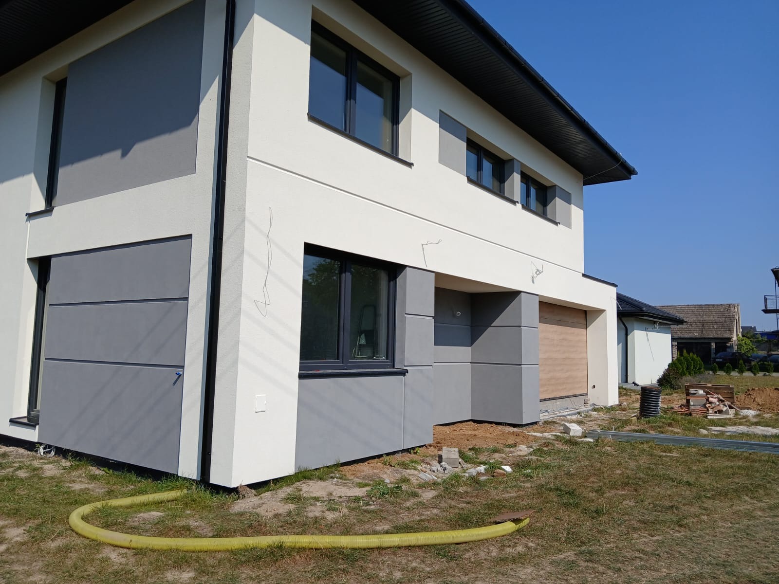 Dwukondygnacyjny dom z elewacją w odcieniach bieli i szarości, widoczne ślady po demontażu rusztowania, żółty wąż na trawniku.