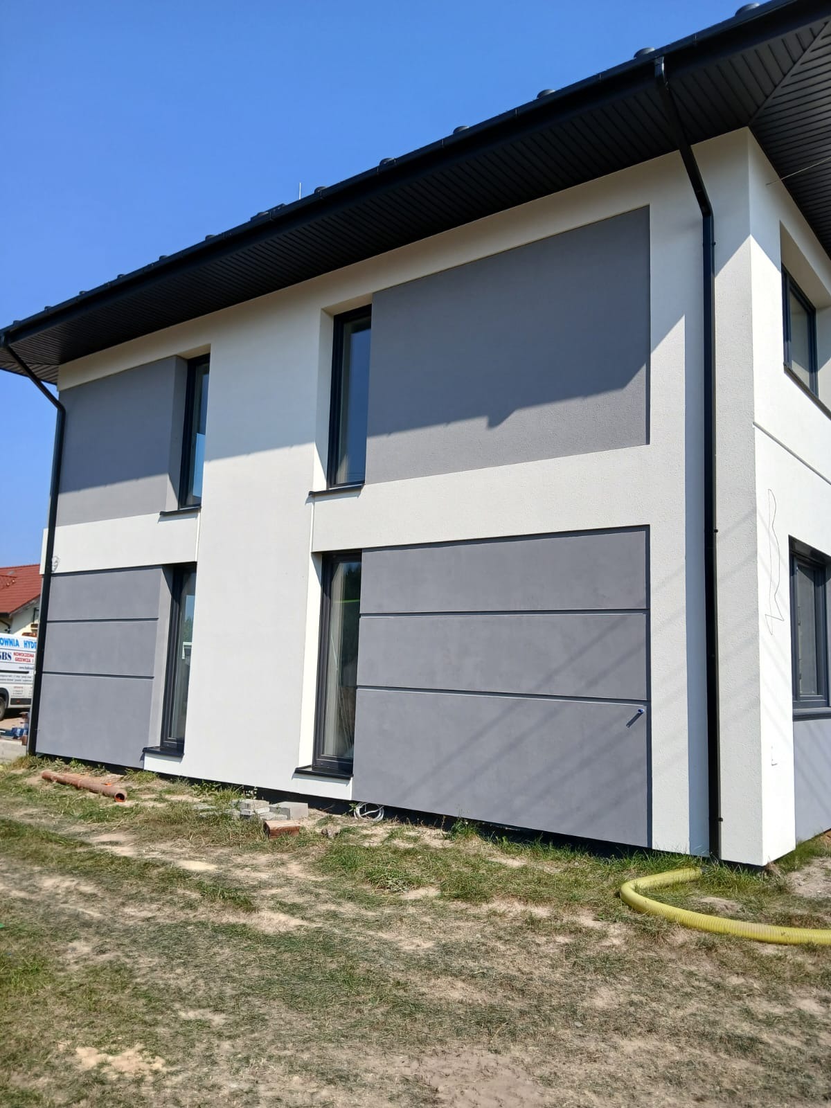 Dwukondygnacyjny dom z nową, dwukolorową elewacją w odcieniach bieli i szarości z widocznymi panelami, czarną stolarką okienną i orynnowaniem, na trawniku leży żółty wąż.