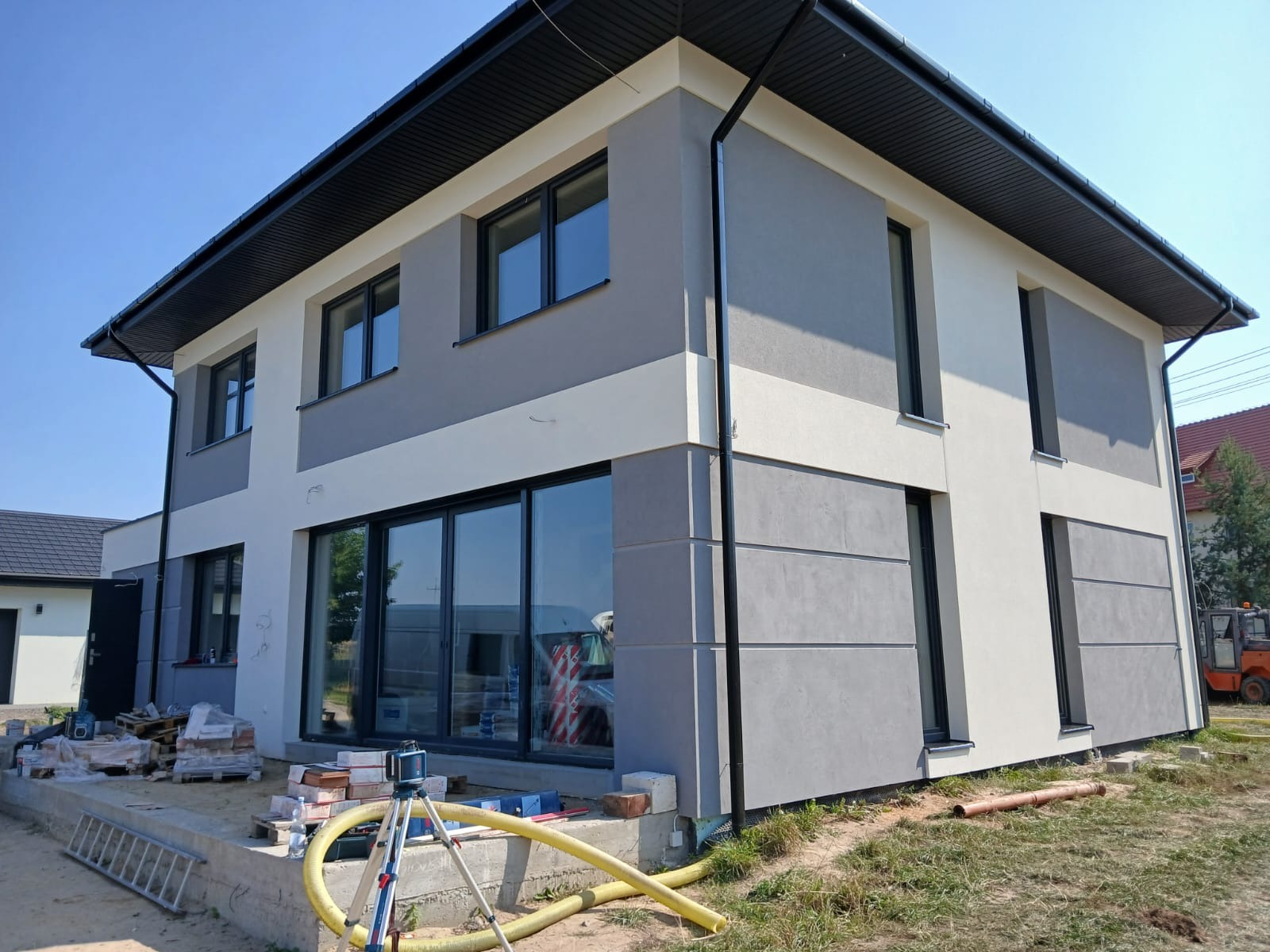 Dwukondygnacyjny dom z nową, dwukolorową elewacją w odcieniach szarości i bieli, widoczne narzędzia budowlane i materiały na placu budowy.