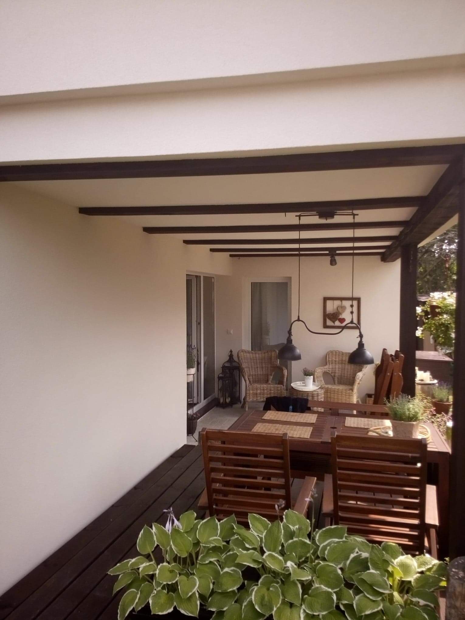 Zadaszony taras z drewnianą podłogą i meblami ogrodowymi, ściana pomalowana na jasny kolor z ciemnymi belkami stropowymi, ozdobne lampy wiszące.