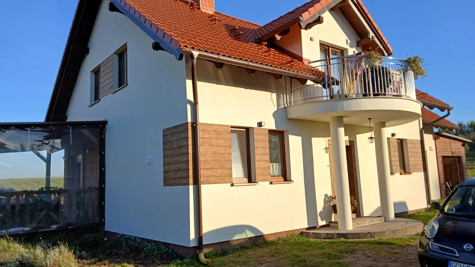 Jasna elewacja domu jednorodzinnego z drewnianymi okiennicami, balkonem i zadaszeniem tarasu z przezroczystymi ścianami.