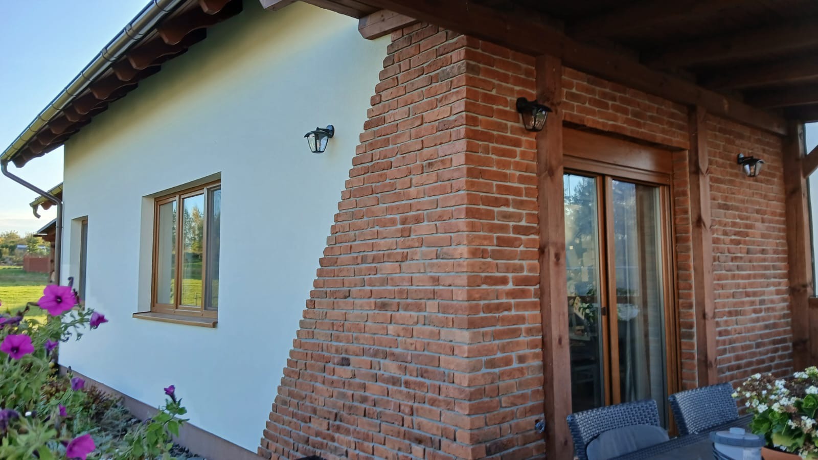 Dom z fragmentem elewacji pokrytej jasnym tynkiem i narożnikiem wykończonym czerwoną cegłą, oświetlonymi przez zewnętrzne lampy.