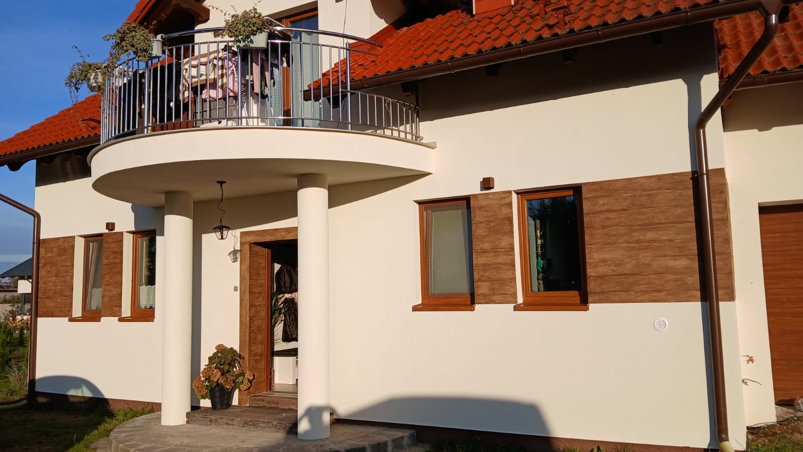 Jasna elewacja domu z brązowymi akcentami imitującymi drewno, balkon z metalową balustradą, czerwona dachówka, widoczne okna z brązowymi ramami.
