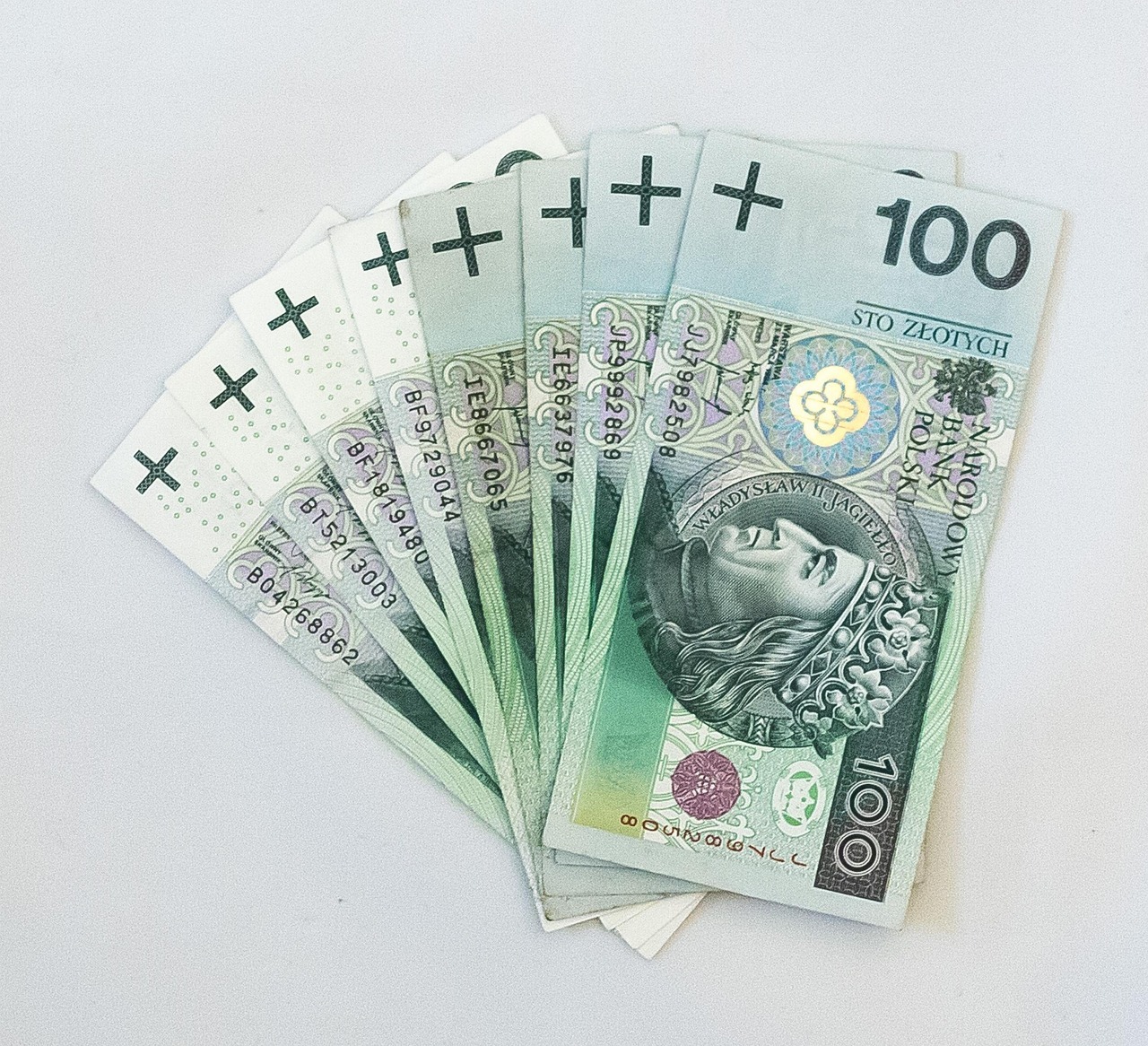 Rozłożony wachlarz banknotów 100 zł na białym tle. Widoczne numery seryjne, wizerunek Władysława Jagiełły i nominał. Symbolizuje finanse, oszczędności lub zysk.